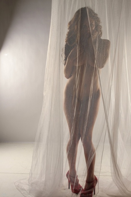 Le modèle amateur Lily Xo pose nu derrière des rideaux transparents en talons hauts