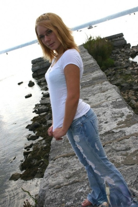 Młoda blondynka w podartych dżinsach zdejmuje koszulkę, aby modelować w staniku nad morzem
