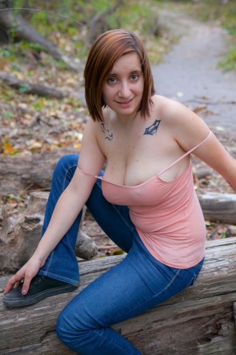 La rossa pallida si sfila i jeans sul sentiero per posare nuda mostrando i tatuaggi