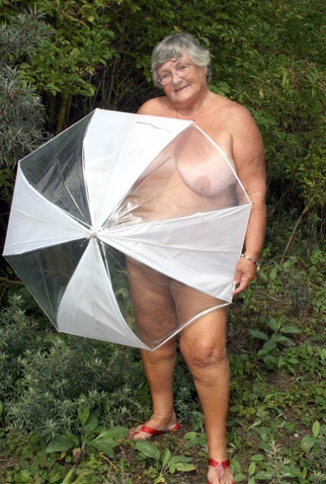 Die fettleibige Oma Libby hält einen Regenschirm und posiert nackt vor Tannenbäumen