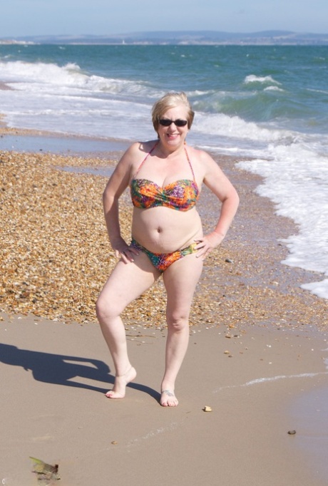 Den modne britiske kvinnen Speedy Bee bærer solbriller mens hun kler seg naken på stranden.