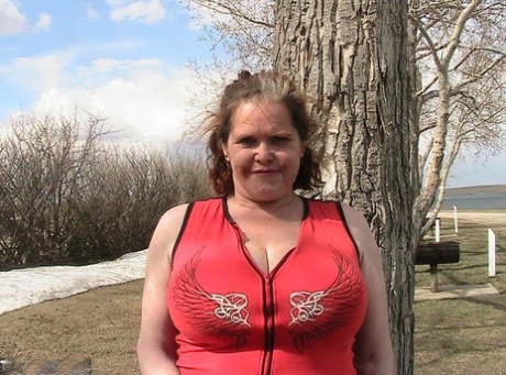 Older redhead Misha MILF displays her large tits by a lake before dildo riding