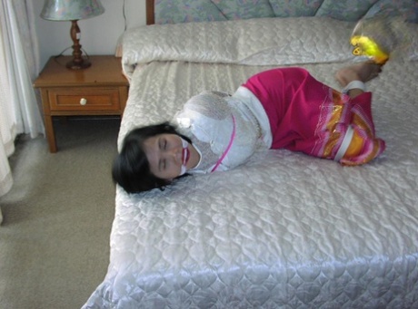 Asiatisches Mädchen wird gespalten und geknebelt, während sie in ihrer Kleidung auf einem Bett gefesselt ist