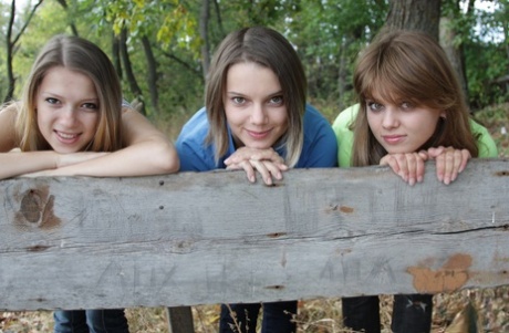 Drie jong uitziende meisjes zitten naakt op een houten bank op het platteland