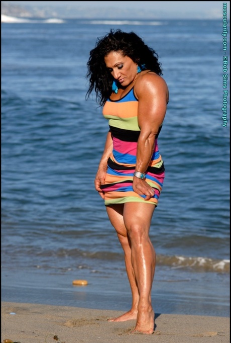 Brunett bodybuilder Tonia Moore flexar sina muskler på en strand i en klänning