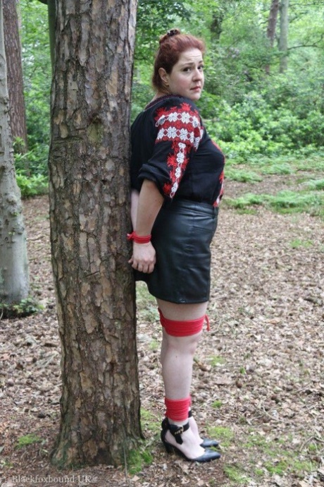 Une rousse épaisse est bâillonnée et attachée à un arbre dans une forêt.