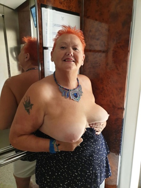 Une grosse femme aux cheveux roux courts appuie ses gros seins et ses fesses contre une vitre.