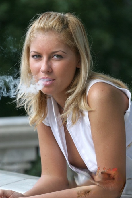 Une blonde naturelle expose ses seins de côté en fumant une cigarette sur un balcon.