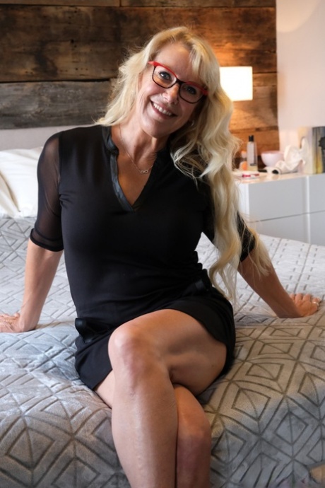 Rondborstige blondine doet haar kleren en bril af voor de seks op een bed