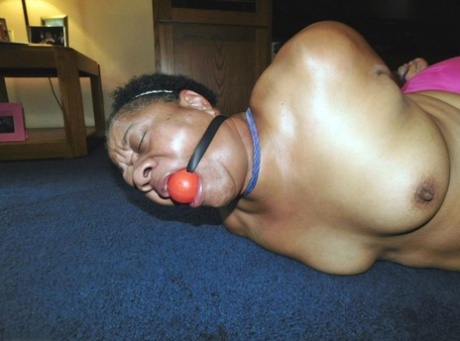 Trixie, een zwarte vrouw met overgewicht, worstelt tegen een ball gag terwijl ze vastgebonden is