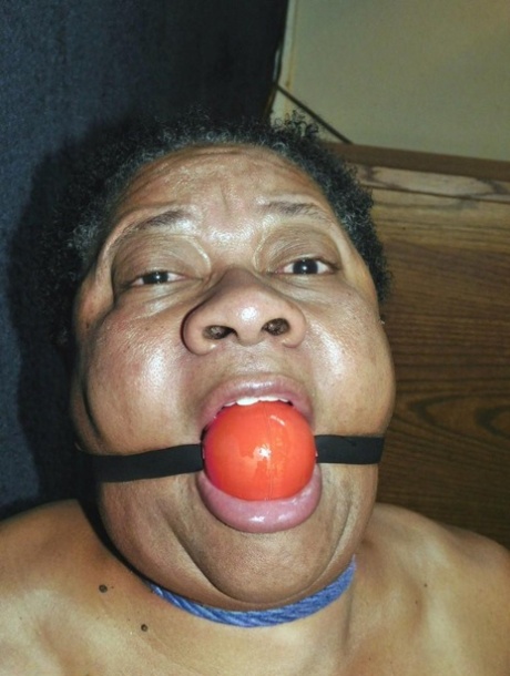 Trixie, een zwarte vrouw met overgewicht, worstelt tegen een ball gag terwijl ze vastgebonden is