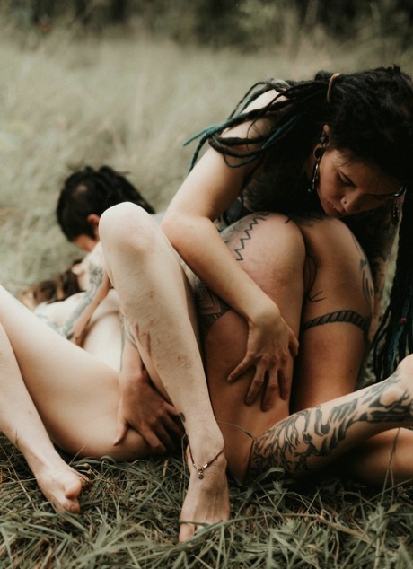 Three tattooed girls share lesbian kisses in long grass near a fir tree