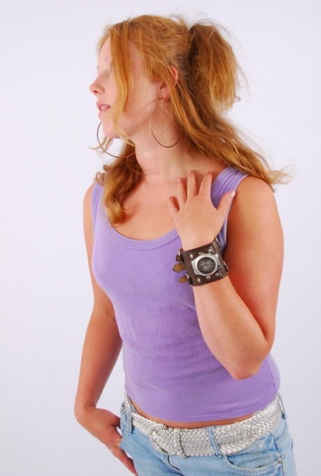 A ruiva natural modela um relógio de punho grande com uma camisola de alças e calças de ganga desbotadas