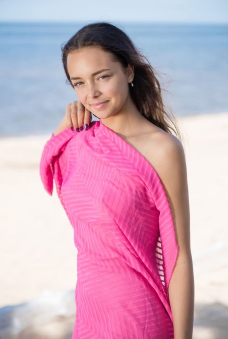 Sweet brunette teen Slava A holds pink fabric while posing nude on a beach