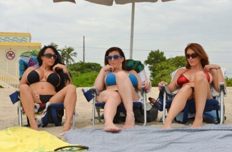 Três mulheres de praia peitudas tiram os tops dos biquínis para exibir as mamas grandes junto à água